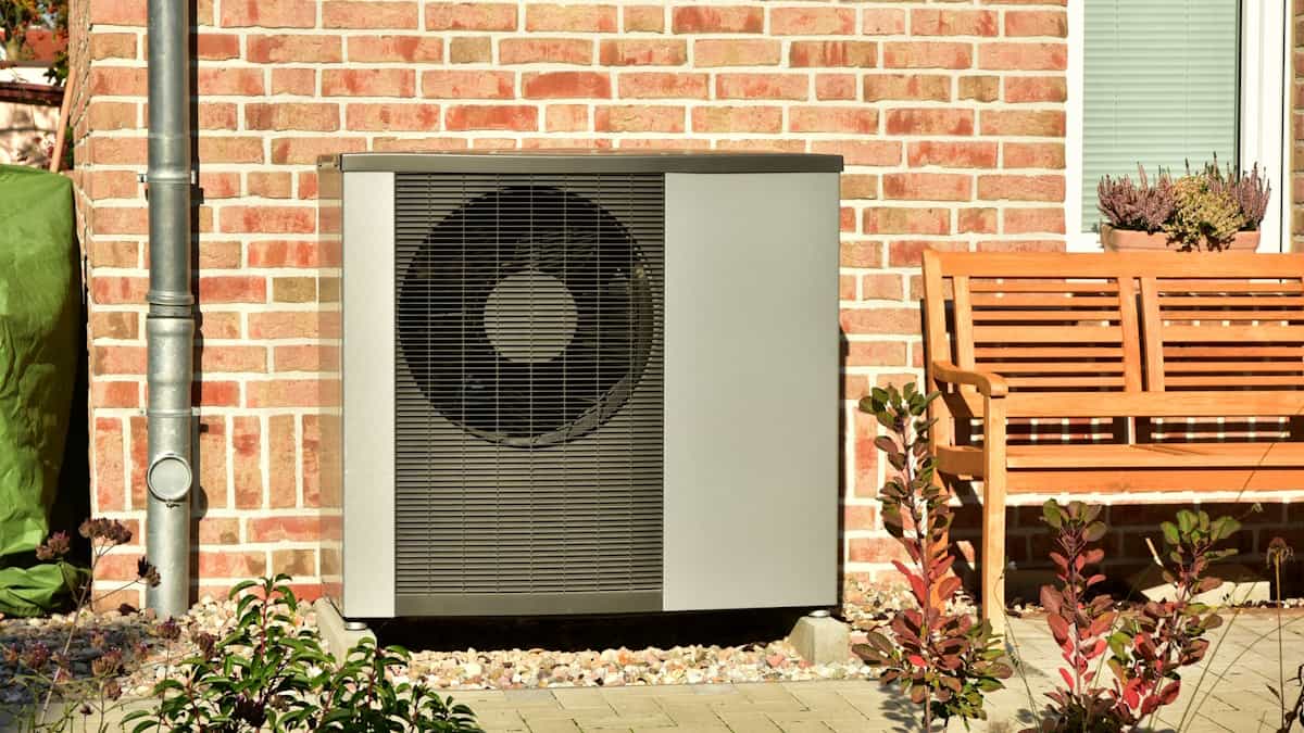 Een warmtepomp is tegen de bakstenen gevel van een woning geplaatst. Rechts een houten bankje, op de voor- en achtergrond planten