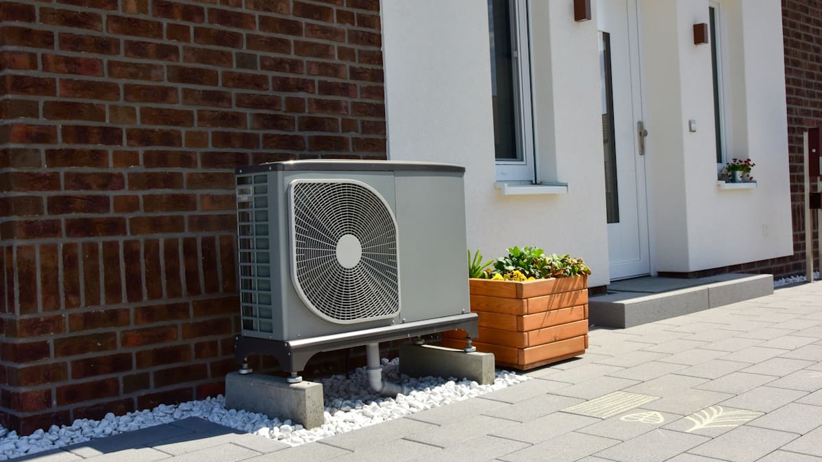 Een grijze luchtwarmtepomp staat waterpas naast de bakstenen gevel van een huis